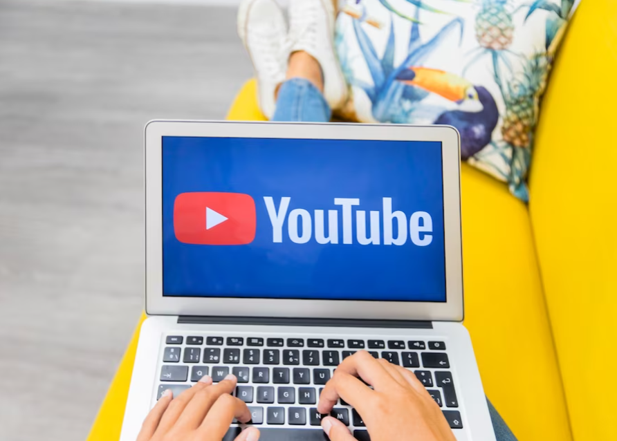 woman on the yellow coach with hands on laptop keyboard and youtube icon on the screen