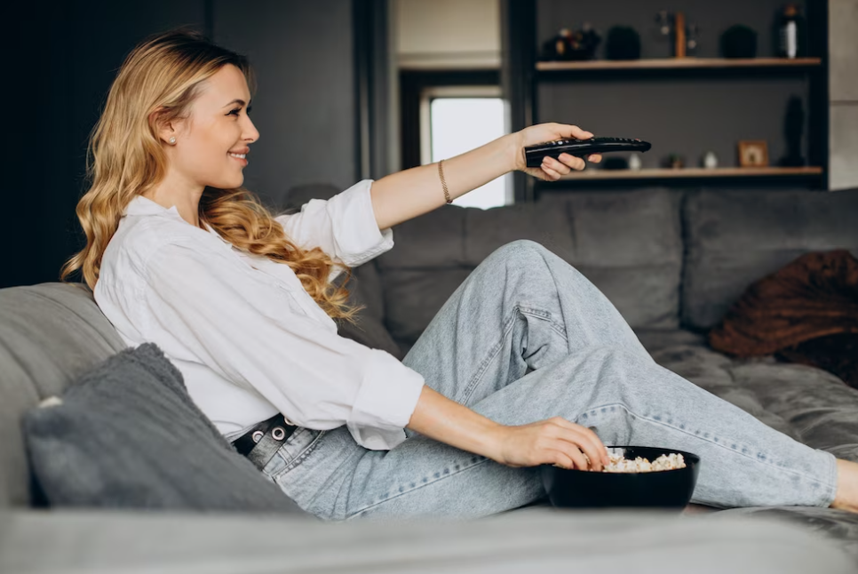 woman in white shirt an jeans sitting on the coach with remote control of TV