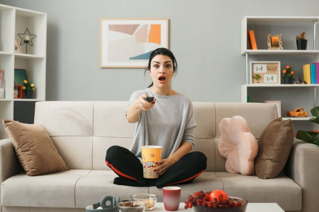 Girl with a bucket of popcorn holding a TV remote control sitting on the sofa